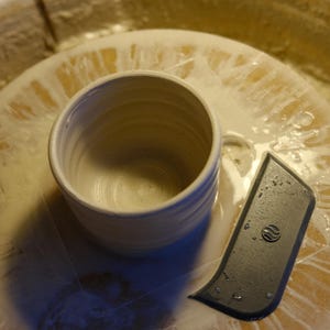 May include: A handmade, off-white ceramic mug with a rounded body and a textured exterior. The mug sits on a pottery wheel next to a black trimming tool. The wheel is covered in a wet, creamy substance.