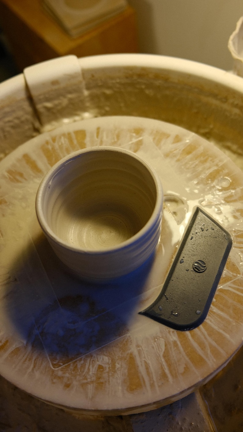 May include: A handmade, off-white ceramic mug with a rounded body and a textured exterior. The mug sits on a pottery wheel next to a black trimming tool. The wheel is covered in a wet, creamy substance.