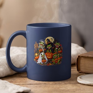 May include: A dark blue ceramic mug featuring a colorful graphic of five cartoon frogs dressed in Halloween costumes, including a mummy, a witch, and a ghost, with pumpkins and a sign that reads Trick or Treat against a full moon background.