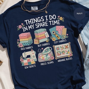 以下が含まれることがあります： THINGS I DO IN MY SPARE TIMEというテキストがプリントされたネイビーブルーのTシャツ。デザインには、積み重ねられた布、ロータリーカッター、ミシン、アイロン、キルトブロックのフレームのイラストが描かれており、それぞれの作業内容を示すラベルが添えられています。