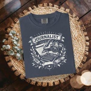 May include: A navy blue t-shirt with a white graphic design featuring the word "JOURNALIST" on a banner. The design includes a pen, camera, typewriter, microphone, and notepad, surrounded by laurel leaves.
