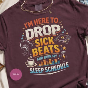 May include: Berry-coloured t-shirt with the text "I'M HERE TO DROP SICK BEATS AND RUIN MY SLEEP SCHEDULE." The graphic includes a turntable, headphones, a coffee cup, and a can with a lightning bolt.