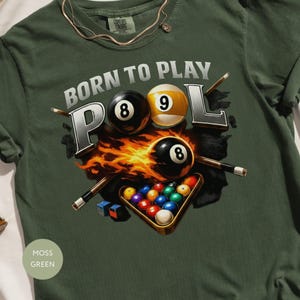 May include: A moss green t-shirt with a pool-themed graphic. The design includes the text "BORN TO PLAY POOL", pool balls with the numbers 8 and 9, flames, pool cues, and a rack of balls.