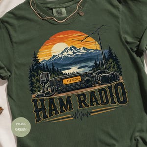 May include: A moss green t-shirt featuring a graphic of a ham radio transceiver, a microphone, and headphones set against a mountain landscape with a sunset. The text HAM RADIO is printed in large dark letters below the equipment illustration.