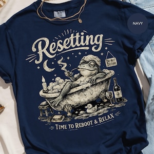 Pode incluir: Camiseta azul marinho com um desenho de um sapo numa banheira, fumando um cachimbo. As palavras "Resetting" e "Time to Reboot & Relax" estão em estilo vintage. Uma placa de "Do Not Disturb" também é visível.