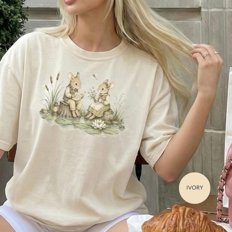 Two bunnies read and hold flowers by a pond on an ivory t-shirt.