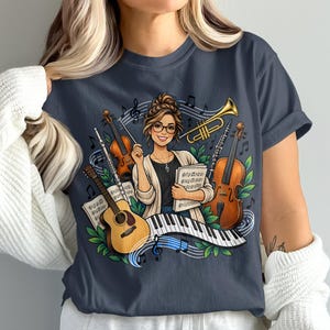 Puede incluir: Una camiseta gris oscuro con una ilustración gráfica de una persona sosteniendo partituras, rodeada por una guitarra, un violín, una trompeta y teclas de piano. Notas musicales y hojas verdes se intercalan en el diseño colorido.