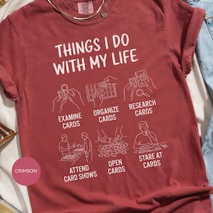 Peut inclure: T-shirt de couleur cramoisie avec l'inscription "THINGS I DO WITH MY LIFE" et des illustrations liées aux cartes. Le design comprend les phrases "EXAMINE CARDS", "ORGANIZE CARDS", "RESEARCH CARDS", "ATTEND CARD SHOWS", "OPEN CARDS" et "STARE AT CARDS".