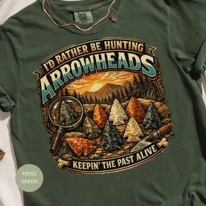 May include: A moss green t-shirt featuring a graphic print with the text I'D RATHER BE HUNTING ARROWHEADS and KEEPING THE PAST ALIVE. The design shows a landscape with mountains, a magnifying glass, and several stone arrowheads on the ground.