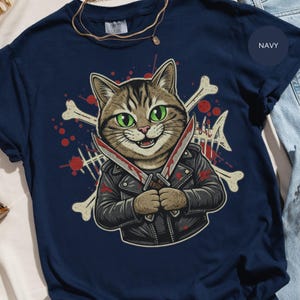 May include: Navy blue t-shirt featuring a cartoon cat with green eyes, wearing a black leather jacket and holding two bloody knives. The design includes crossbones and red blood splatters. The shirt has the words "Comfort Colors" printed on the top.