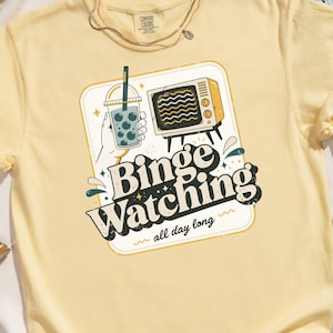 May include: Yellow Comfort Colors t-shirt with a graphic that says "Binge Watching all day long." The graphic includes a bubble tea, a vintage television, and a hand holding the bubble tea.