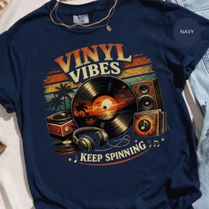 Puede incluir: Camiseta azul marino con un diseño gráfico retro. El diseño presenta las palabras "VINYL VIBES" sobre un disco de vinilo, palmeras y una escena de puesta de sol. La frase "KEEP SPINNING" está debajo, con notas musicales.