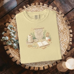 May include: A light yellow Comfort Colors t-shirt featuring a watercolor illustration of macrame supplies. The design includes a macrame wall hanging, a spool of thread, scissors, and an open book. The shirt is displayed on a woven mat.