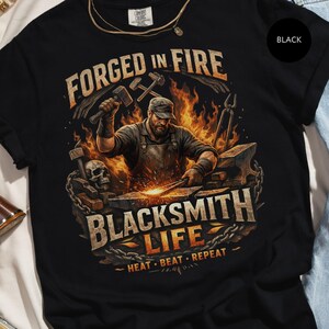 May include: Black t-shirt with a blacksmith graphic. The design features a blacksmith working with fire and tools, with the words "FORGED IN FIRE" and "BLACKSMITH LIFE" prominently displayed. The phrase "HEAT. BEAT. REPEAT." is also included.