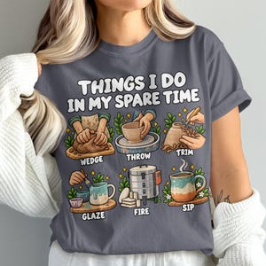 May include: A charcoal gray t-shirt featuring the text "THINGS I DO IN MY SPARE TIME" above six illustrations of pottery activities: wedge, throw, trim, glaze, fire, and sip, each with corresponding graphics of clay, tools, and a kiln.