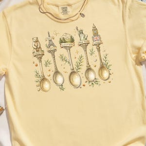 May include: A light yellow t-shirt with a graphic of decorative spoons, each topped with a landmark: a bear for California, the Eiffel Tower for Paris, mountains for Colorado, the Statue of Liberty for New York, and Big Ben for London.