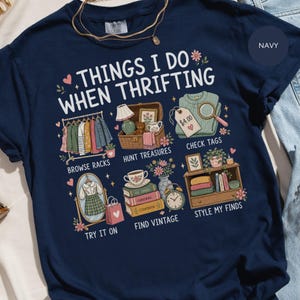 Pode incluir: Uma t-shirt azul-marinho com o texto "THINGS I DO WHEN THRIFTING" e ilustrações de araras de roupas, uma cesta de tesouros, uma etiqueta de preço, um espelho com um vestido, livros vintage e uma prateleira de madeira com decoração.