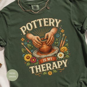 May include: Moss green t-shirt with a pottery design. The graphic features hands shaping a clay pot, surrounded by flowers and paintbrushes. Text reads "POTTERY IS MY THERAPY" in a vintage style.
