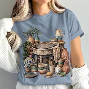 May include: A light blue t-shirt featuring a graphic illustration of a pottery studio scene. The design displays a central pottery wheel surrounded by various ceramic vessels, including vases, bowls, and pitchers, along with sculpting tools.
