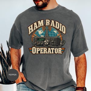 May include: A charcoal grey t-shirt featuring a graphic print of a radio transceiver, a globe, and a transmission tower. The text reads HAM RADIO OPERATOR. The design includes orange, blue, and cream colours, with a vintage-inspired aesthetic.