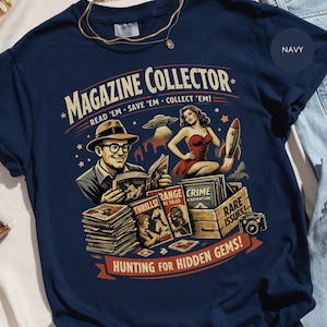 May include: Navy blue t-shirt featuring a vintage-style illustration. The design includes the text "MAGAZINE COLLECTOR" with a retro graphic of a man reading magazines, a woman, a rocket, and the text "HUNTING FOR HIDDEN GEMS!"