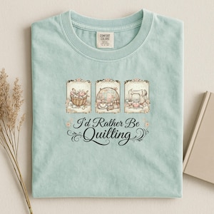 May include: A pale blue t-shirt with the phrase "I'd Rather Be Quilting." The design includes three framed images: a basket of sewing supplies, a tea set, and a sewing machine, all in a vintage style.
