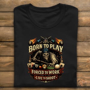 Op de afbeelding: Zwart t-shirt met een biljartthema. Het ontwerp toont een schedel die aan het poolen is, vergezeld van de tekst "BORN TO PLAY, FORCED TO WORK, LIVE TO SHOOT." Inclusief poolballen, speelkaarten en vlammen.