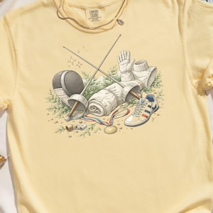 May include: A light yellow t-shirt featuring a watercolor illustration of fencing equipment. The design includes a mask, gloves, foil, and a sneaker. A gold medal and the text "COMFORT COLORS" are also visible.