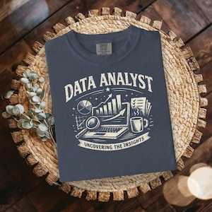 Puede incluir: Una camiseta azul marino con un gráfico blanco que dice "DATA ANALYST" encima de un portátil, una lupa, un gráfico circular y una taza de café. El texto "UNCOVERING THE INSIGHTS" está debajo. La camiseta se muestra sobre una estera tejida.