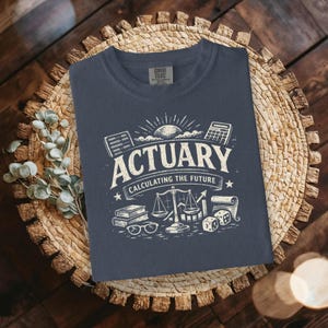 May include: A navy blue t-shirt featuring a white graphic with the words "ACTUARY CALCULATING THE FUTURE." The design includes an abacus, calculator, scales, books, glasses, dice, and a scroll. The shirt is displayed on a woven surface.