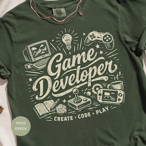 以下が含まれることがあります： 「Game Developer」および「CREATE CODE PLAY」という文字が記されたクリーム色のグラフィックが特徴のモスグリーンのTシャツ。デザインには、コンピューター、ゲームコントローラー、電球、ジョイスティック、携帯ゲーム機、ノートのイラストが含まれています。