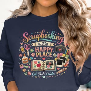 May include: Navy blue sweatshirt with a colorful graphic that reads "Scrapbooking is My Happy Place." The design includes scissors, glue, a camera, and an open scrapbook with hearts and flowers. The phrase "Cut, Paste, Create!" is also included.