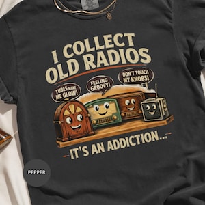 Pode incluir: Camiseta cinza escuro com o texto "I Collect Old Radios" e rádios de desenho animado. A camiseta apresenta as frases "Tubes Make Me Glow!", "Feeling Groovy!" e "Don't Touch My Knobs!". A frase "It's an Addiction..." também está incluída.