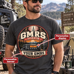 May include: A charcoal gray t-shirt featuring a circular graphic with a radio tower, a mountain landscape, and a two-way radio. The text reads GMRS OPERATOR and YOUR GMRS. The design includes red and cream accents, with arrows pointing to areas for personalization.