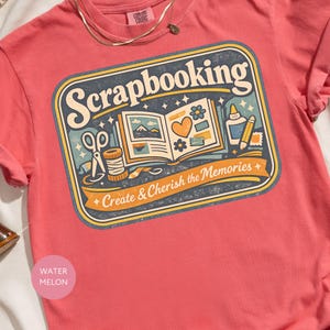May include: A coral-colored t-shirt with a graphic that reads "Scrapbooking" in a vintage style. The design includes scissors, a spool of thread, an open scrapbook, and the text "Create & Cherish the Memories."