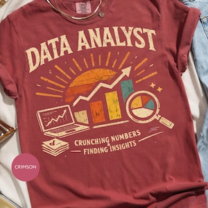 May include: A crimson short-sleeve t-shirt featuring the text DATA ANALYST in a curved font above a graphic of a laptop, a rising bar chart, and a magnifying glass over a pie chart. Below the graphic is the text CRUNCHING NUMBERS & FINDING INSIGHTS.