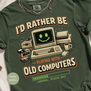 以下が含まれることがあります： モスグリーンのTシャツ。テキストは「I'd Rather Be Playing With Old Computers」です。グラフィックは、笑顔の画面、キーボード、その他のヴィンテージ技術を備えたレトロなコンピューターセットアップです。シャツは柔らかい素材で作られています。