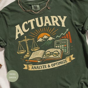 Puede incluir: Una camiseta verde musgo con un gráfico que incluye el texto ACTUARY y ANALYZE & OPTIMIZE. El diseño muestra una balanza de justicia, un libro abierto con gafas, una calculadora, un gráfico de barras ascendente y un sol sobre montañas.