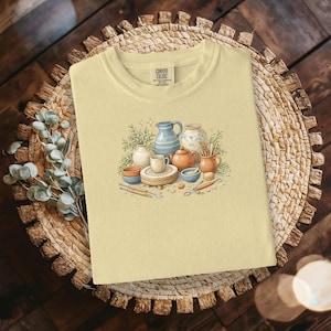 May include: A pale yellow Comfort Colors t-shirt with a watercolour illustration of pottery tools and ceramic items, including vases, bowls, and a teapot. The design features a pottery wheel, brushes, and a mug. The shirt is on a woven mat.