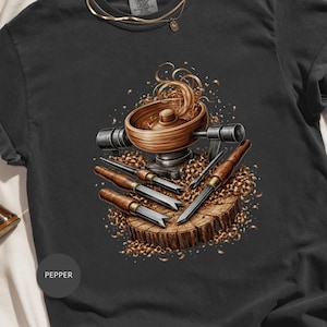 May include: Dark gray t-shirt featuring a detailed illustration of woodworking tools and a wooden bowl on a tree stump. The design includes wood shavings and various chisels, showcasing a craft theme.