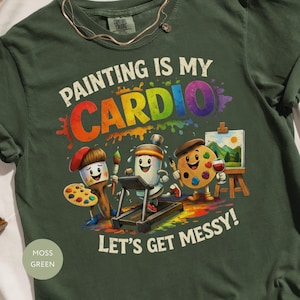 Puede incluir: Una camiseta verde musgo con un gráfico que incluye el texto "PAINTING IS MY CARDIO" y "LET'S GET MESSY!". El diseño muestra personajes de dibujos animados, incluyendo un pincel, un tubo de pintura en una cinta de correr y una paleta.