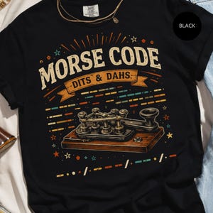 May include: Black t-shirt with a vintage-style graphic featuring the words "MORSE CODE" and "DITS & DAHS." The design includes a Morse code key and decorative elements in orange, blue, and green. The shirt is made of a soft material.