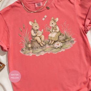 Two watercolor mice sit by a pond, one reading a book, the other holding flowers.