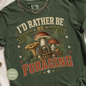May include: A moss green t-shirt featuring a graphic print of various mushrooms, a woven basket, and fern leaves. The text "I'D RATHER BE FORAGING" is printed in a bold, distressed font. The design includes small star accents and berries.