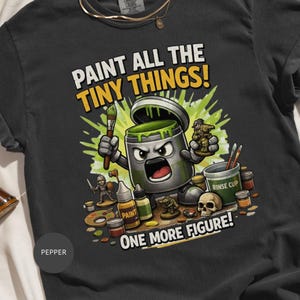 May include: A charcoal grey t-shirt featuring a graphic of an anthropomorphic paint pot holding a brush and a miniature figure. The text reads PAINT ALL THE TINY THINGS! and ONE MORE FIGURE!. Surrounding elements include paint bottles, a skull, and a rinse cup.