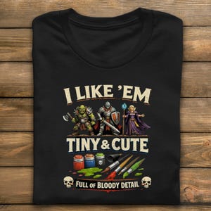 May include: Black t-shirt with the text "I LIKE 'EM TINY & CUTE FULL OF BLOODY DETAIL". The shirt features illustrations of fantasy characters, paint pots, brushes, and skulls.
