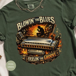 May include: A moss green t-shirt with a musical design. The graphic includes a harmonica, guitar, and a musician's silhouette. Text reads "BLOWIN' the BLUES" and "FEELIN' the GROOVE". Musical notes and flames are also included.