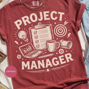 May include: Crimson t-shirt featuring the text "PROJECT MANAGER" in white, accompanied by illustrations of a lightbulb, laptop, clipboard, coffee mug, and target. The shirt has a crew neck and short sleeves.