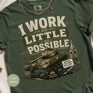 May include: A moss green t-shirt featuring the text I WORK AS LITTLE AS POSSIBLE. The graphic includes a 1/35 scale model tank, a bottle of olive drab paint, a bottle of plastic model cement, a hobby knife, and a sign reading BECAUSE IT'S A HOBBY, NOT A JOB.