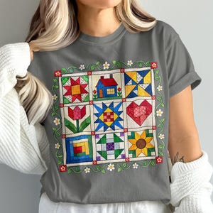 May include: A charcoal grey t-shirt featuring a central graphic of a nine-patch quilt design. The quilt squares display colourful patterns including a red star, a blue house, a tulip, a heart, and a sunflower, all framed by a green vine with flowers.
